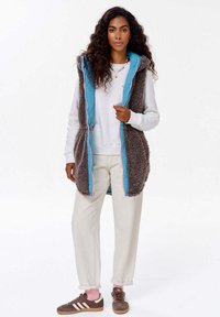 Šedá chlupatá vesta s modrou podšívkou, zipem vpředu a růžovými akcenty. Nosič ji má přes bílou mikinu a sladěnou s krémovými kalhotami a hnědými teniskami.