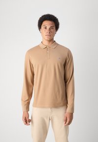 Camisa polo de manga larga en color beige con cuello y dos botones. Presenta un pequeño logo azul en el lado izquierdo del pecho. Textura suave y corte relajado.