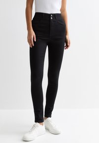 Jean skinny noirs taille haute avec poches avant, fermeture à deux boutons et une texture denim classique lisse. Portés avec des baskets blanches.