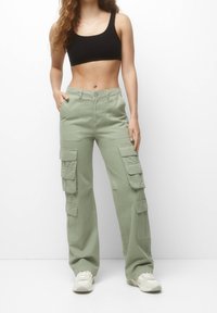 Kvinde iført salviegrønne cargobukser, sort crop top og hvide sneakers, stående foran en ensfarvet hvid baggrund.