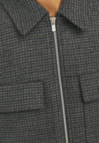 Veste en laine grise avec un motif à carreaux texturé, dotée d'une fermeture éclair argentée et de deux poches avant avec rabats. Design avec col.