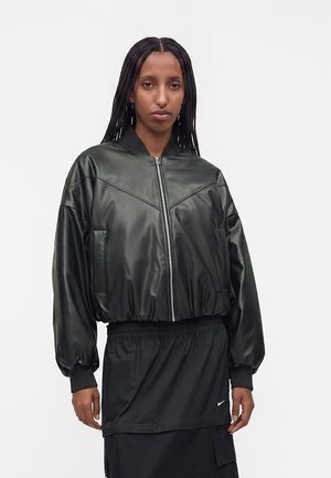 Femme aux longs cheveux tressés portant une veste en cuir noire zippée et une jupe noire avec un petit logo blanc sur un fond clair uni.