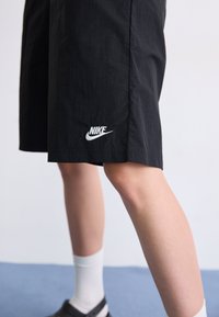 Person trägt schwarze Nike-Shorts, weiße Kniestrümpfe und schwarze Slip-On-Schuhe, steht auf blauem Boden vor weißer Wand.