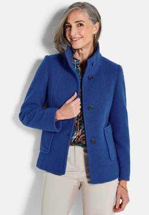 Veste en mélange de laine bleu avec un col haut, tissu texturé, deux poches avant et fermeture à boutons. Portée par-dessus un haut à motifs.
