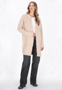Langer beiger Cardigan aus flauschigem Material, mit rundem Halsauschnitt, Knopfverschluss und gerippten Bündchen, kombiniert mit schwarzen Jeans.
