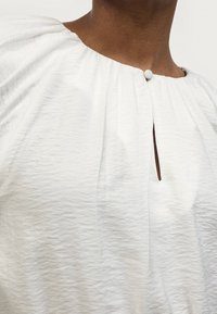 Chemisier blanc texturé avec un col rond, présentant de discrets froncés et une petite ouverture en forme de clé fermée par un bouton assorti.