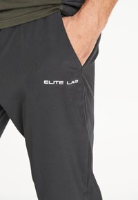 Schwarze Sporthosen aus leichtem, glattem Stoff mit seitlichen Taschen und einem weißen "ELITE LAB"-Logo.