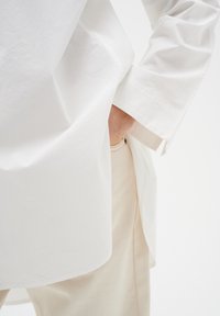 Balta garām piedurknēm krekls ar brīvu piegriezumu, ar gludu tekstūru un sānu spraugām, apvienots ar gaiši bēšiem biksēm ar kabatas detaļu.