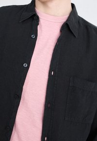 Svart skjorta med krage, med en framficka och blå knappar, som är lager-på-lager över en ljusrosa t-shirt med rund hals.