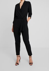 Mono negro ajustado con un escote en V profundo, mangas 3/4 y cinturilla atada. Presenta piernas entalladas y una textura suave; se lleva con tacones negros con tiras.