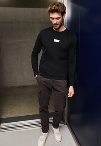Black long-sleeve shirt with a small white label, paired with dark cargo pants featuring side pockets and white sneakers. Casual attire.