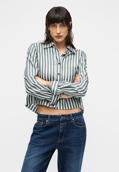 Πουκάμισο με πράσινες και λευκές ρίγες, κουμπωτό με μακριά μανίκια, συνδυασμένο με μπλε τζιν. Το πουκάμισο έχει cropped σχεδιασμό και λεπτομέρεια τσέπης.