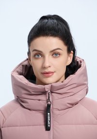 Lichtroze pufferjack met een hoge kraag, voorzien van quiltdetails en een ritsluiting. Zwarte trekproef met logo benadrukt het ontwerp.