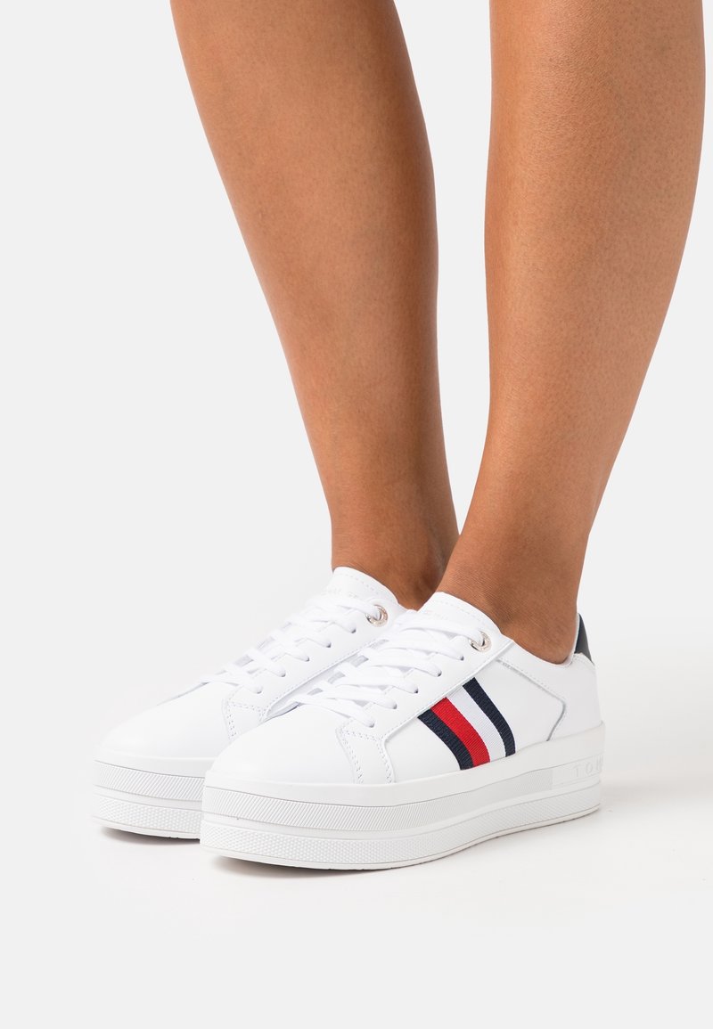 Weiße Schnürsenkel-Sneaker mit einer strukturierten Gummisohle, die an der Seite einen blau-roten Streifenakzent und metallische Ösen aufweist.