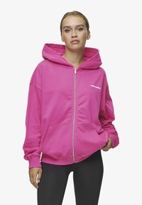 Roza zip-up majica sa velikom kapuljačom, prednjim dzepovima i rebrastim manžetama. Ima bijeli logo tekst na lijevoj strani prsnog područja.