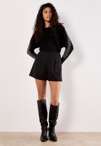 Frau mit langen lockigen Haaren trägt einen schwarzen Pullover mit weißen Perlendetails, schwarze hoch taillierte Shorts und knielange schwarze Lederstiefel.