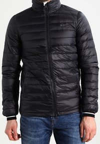 Svart pufferjacka med quiltat mönster, hög krage, hel dragkedja och sidofickor. Har ribbstickade muddar och en slät, glänsande yta.