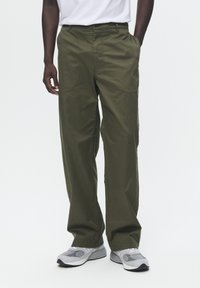 Olive green, relaxed-fit pants made of soft fabric with side pockets, featuring a button and zipper closure, worn with gray sneakers.