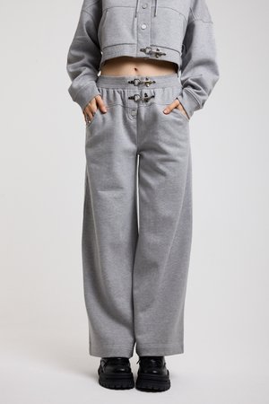 Femme portant un pantalon de survêtement gris à jambes larges et un sweat à capuche court assorti avec des détails en métal, mains dans les poches, et chaussures noires épaisses.
