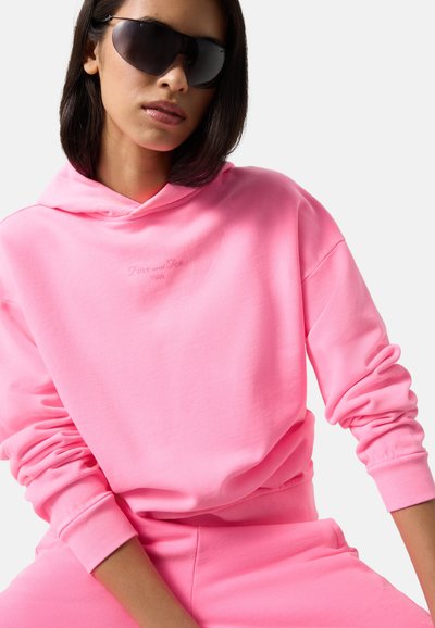 Femme portant un sweat à capuche rose vif oversize et un pantalon assorti avec de grandes lunettes de soleil sombres sur un fond uni.
