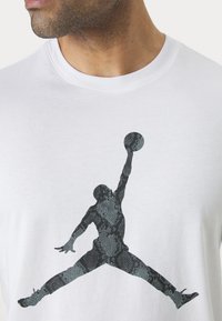 Hvid bomulds T-shirt med en stor sort silhuet af en basketballspiller med et slangemønster, placeret mod en ensfarvet baggrund.
