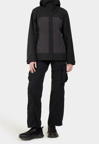 Veste noire imperméable avec des accents gris, dotée d'une capuche et de deux poches latérales. Associée à un pantalon cargo noir et des baskets noires.