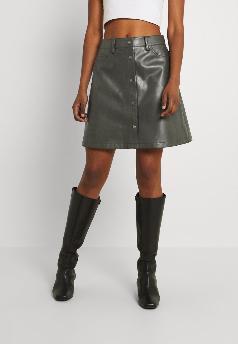 Femme portant une jupe en cuir boutonnée vert foncé, un haut court blanc et des bottes noires jusqu'aux genoux, debout devant un fond clair uni.