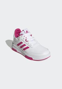 Chaussure de sport blanche avec des accents roses, dotée de trois bandes iconiques, d'un laçage et d'une semelle en caoutchouc texturé pour une meilleure adhérence.