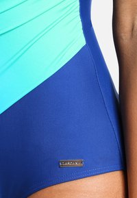 Maillot de bain avec un haut turquoise et un bas bleu marine. Fabriqué en tissu lisse avec un détail de logo métallique sur le côté.