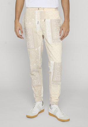 Pantalon de jogging beige slim avec motif cachemire blanc, équipé de poignets élastiques, porté avec des baskets blanches et une chemise blanche.