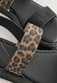 Svarta kilsandaler med en texturerad, vävd sula. Två remmar: en i svart mocka och en i leopardskrutigt tyg med en slät yta.