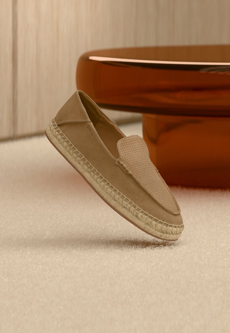 Μπεζ παπούτσι τύπου slip-on με υφή στην πάνω επιφάνεια και υφαντή σόλα, τοποθετημένο όρθιο σε ανοιχτό χαλί δίπλα σε ένα γυαλιστερό καφέ κυκλικό τραπέζι.