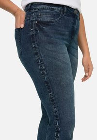 Jeans de mezclilla oscura con un diseño ajustado, que presentan una franja lateral con estampado y un estilo clásico de cinco bolsillos, tela texturizada y herrajes metálicos.