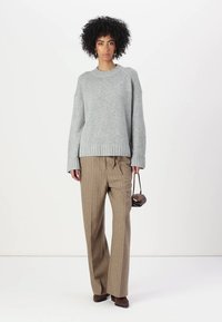 Grauer gestrickter Pullover mit geripptem unteren Saum, kombiniert mit weit geschnittenen braunen Nadelstreifen-Hosen. Das Model hält eine kleine dunkle Handtasche.
