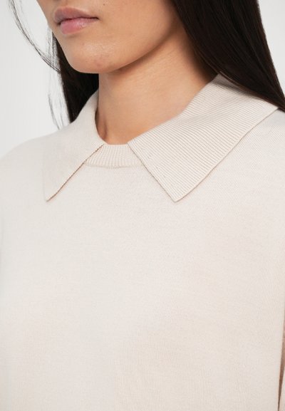 Maglione beige lavorato a maglia con un colletto piatto e testurizzato e dettagli a coste sullo scollo. Il tessuto sembra morbido e liscio.