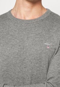 Nahaufnahme einer Person, die einen grauen Strickpullover mit rundem Ausschnitt und kleinem GANT-Logo auf der Brust trägt.