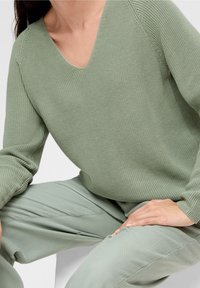 Maglione lavorato a maglia verde chiaro, con scollo a V e maniche lunghe, abbinato a pantaloni leggeri dello stesso colore. I dettagli sono minimalisti e complementari.