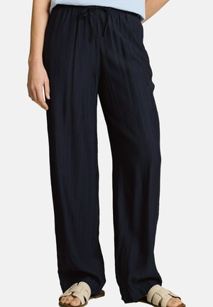 Woman wearing loose black textured pants with an elastic waistband and beige open-toe slide sandals, standing against a white background.
