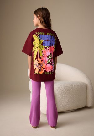 Meisje met de rug naar de camera, draagt een bordeaux oversized T-shirt met tropische print en paarse uitlopende broek, staand bij een witte gebogen stoel.