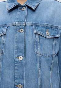 Veste en jean bleu clair avec fermeture boutonnée à l’avant et poches poitrine à rabat, portée par une personne montrant le col et le haut du torse.