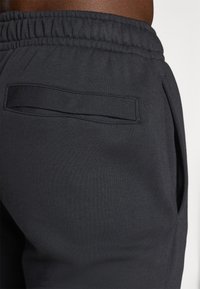 Black athletic joggers featuring a wide elastic waistband, a single back pocket with a flap, smooth texture, and fitted design.