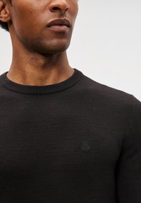 Černý pletený svetr s kulatým výstřihem, texturovaný materiál a malá logo akcentace na hrudi. Jednoduchý, klasický design.