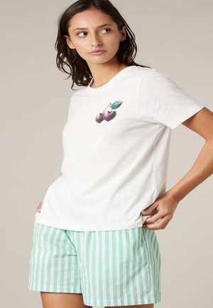 Femme portant un t-shirt blanc avec un motif de cerise et un short à rayures verticales vert menthe et blanc, debout la main sur la hanche devant un fond uni.