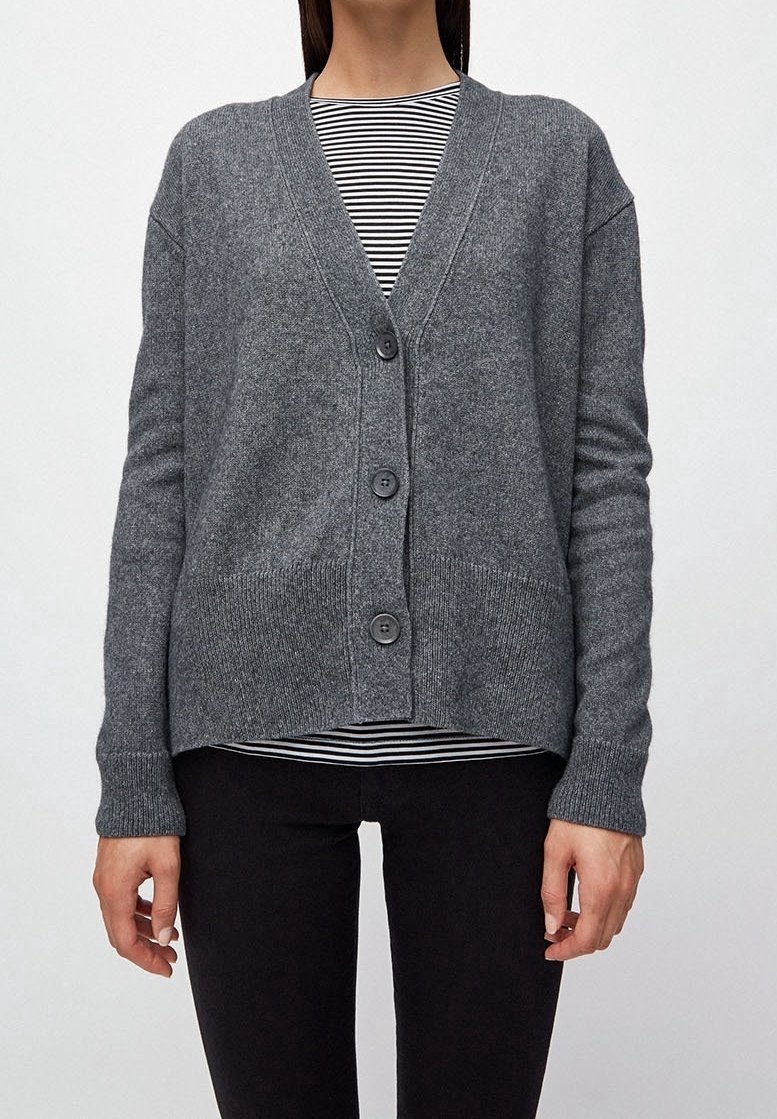 Femme portant un cardigan gris boutonné par-dessus une chemise rayée noir et blanc et un pantalon noir, debout devant un fond uni.
