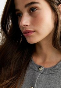 Grå stickad cardigan med rund halsringning, texturerad tyg och en distinkt glansig silverknapp vid stängningen, delvis synlig.