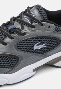 Scarpa sportiva grigia con tomaia in rete, design elegante, lacci neri, accenti blu e un logo prominente sul lato. Suola resistente con texture.