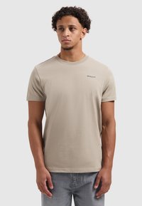 Korte mouwen beige t-shirt van zachte stof. Heeft een ronde halslijn en een klein donker logo op de linkerborst. Model draagt lichte spijkerbroek.