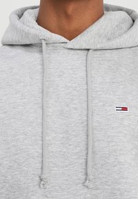 Felpa con cappuccio grigio chiaro dal texture morbido, presenta un cappuccio con coulisse e una piccola etichetta logo in rosso, bianco e blu sul petto.