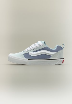Chaussure de skate basse en bleu clair et gris avec lacets blancs, bande latérale blanche, semelle épaisse blanche, et logo rouge sur la languette du talon.