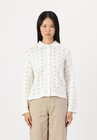 Bela pletena kardigan s čipkastim vzorcem, z gumbi spredaj, rebrastim ovratnikom in manšetami. V kombinaciji z bež hlačami, ki prikazujejo sproščenega obliko.